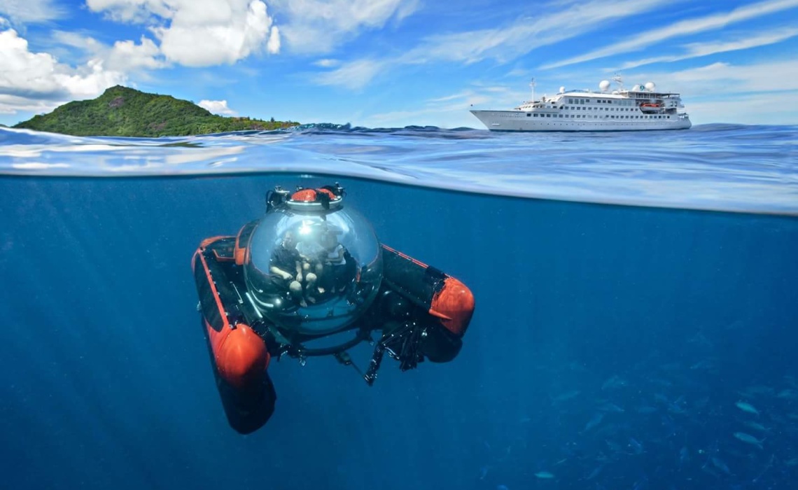 Unterwasserfahrzeug mit gl&auml;serner Kugelkabine taucht im offenen Meer, oberhalb der Wasseroberfl&auml;che ist ein Schiff und eine Insel zu sehen.