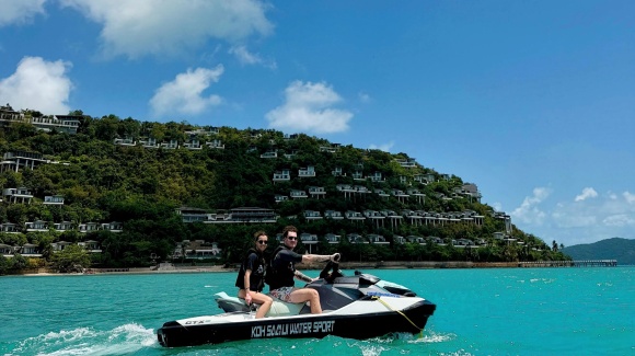 Eine Frau und ein Mann auf einem Jetski, im Hintergrund eine Hotelanlage im Gr&uuml;nen 