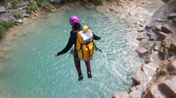 Eine Person springt mit Helm und Rucksack in ein tiefes Wasserloch